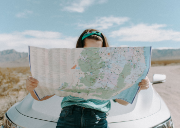 A girl with brown hair sits on the hood of a car while looking at a map