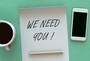 A note in between a cell phone and a cup of coffee that reads, "We need you!"