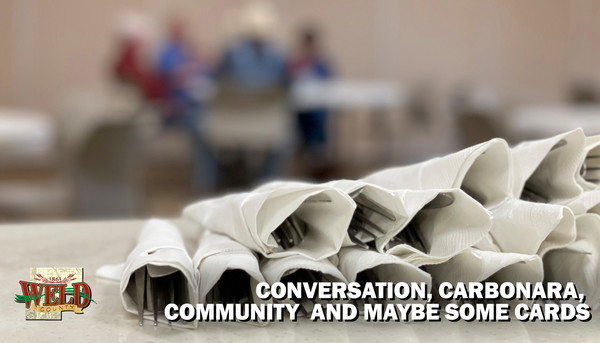 Silverware wrapped in napkins on a table with people faintly in the background. Text reads: Conversation, carbonara, community and maybe some cards.