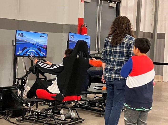 Kids playing a driving video game, in chairs with steering wheels, while a woman and young boy watch