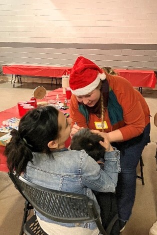 A woman in a Santa hat face-painting a child while sitting on their mother's lap
