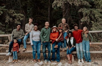 Two adults surrounded by eight children; all are smiling and leaning against a wooden fence beside a forest.