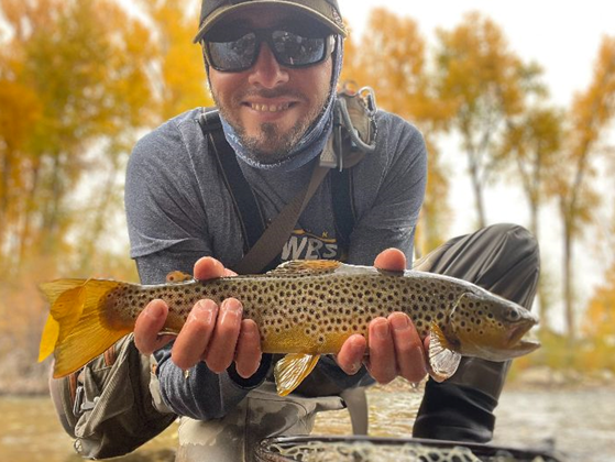 Bobby Lucero, in fishing gear and sunglasses, smiling and showing off a trout he caught.