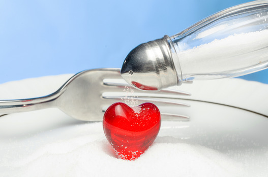 A red plastic heart in a pile of salt. A salt shaker is pouring salt from above and there is a fork in the background.