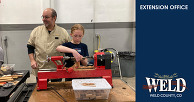 A child working on a woodworking project, while an adult stands in the background.