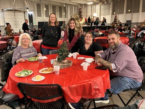 Guests at the holiday Christmas party sitting at a table decorated in holiday decor with plates of salad, pasta and cake.