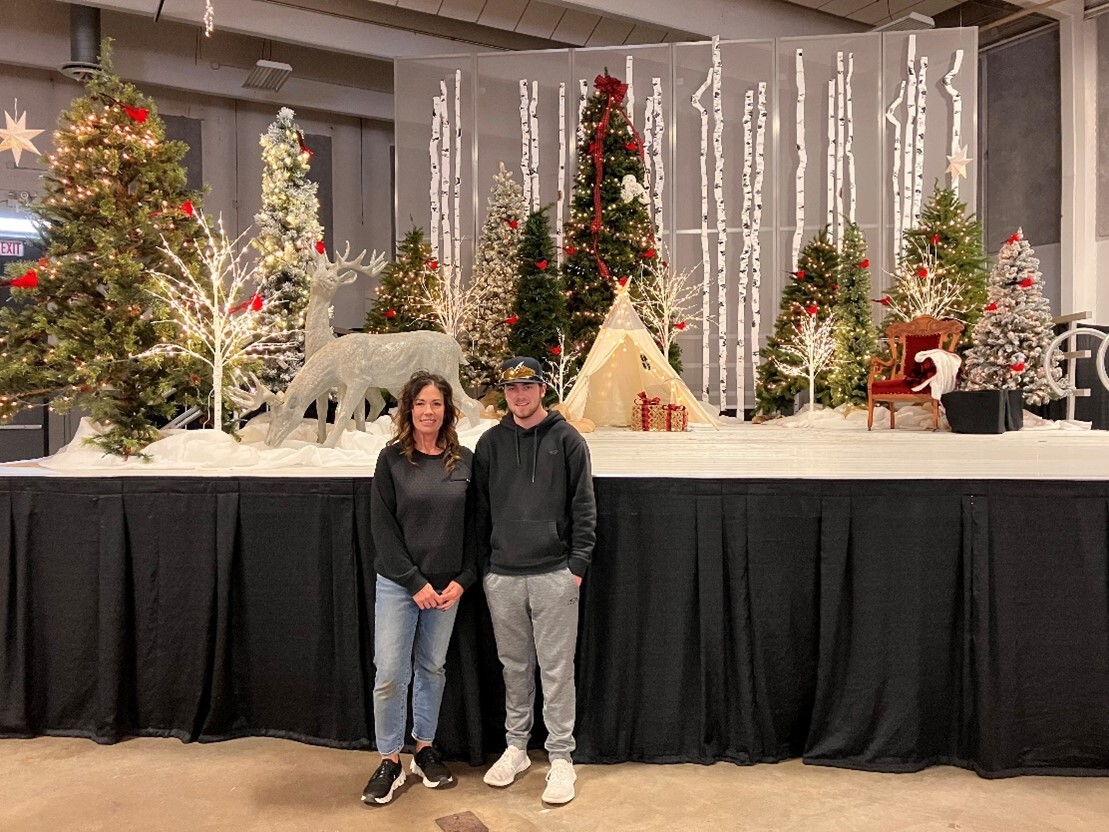 A woman in a black long sleeve top and blue jeans and an adolescent boy in a black long sleeve and grey pants posing in front of a holiday set up.