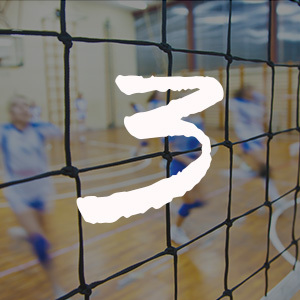 A volleyball net is in focus. In the background is a youth volleyball team practicing.