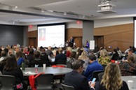 A room full of professionals watching a speaker give a presentation.