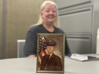 Framed photo of Ron in military uniform on desk. Wanda is in background, out of focus. 