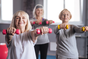Three older women have their arms outstretched while holding dumbbells