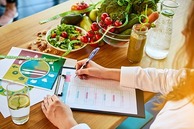 Person filling out a nutrition planner at a table with fresh vegetables, salad, and a smoothie, promoting National Nutrition Month.