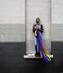 person stands against gray brick wall holding Pride flag
