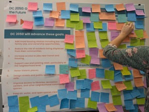 Meeting attendee posts a post-it with a comment on a board listing out the goals for DC 2050
