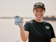 Woman wearing a hat stands smiling holding a piece of trash in her gloved hand in front of a body of water.
