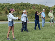 A group of six individuals standing on grass with fishing rods practicing casting.