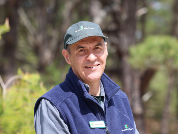 Man wearing a dark green cap and navy fleece vest stands outdoors, smiling, with blurred trees and greenery in the background.