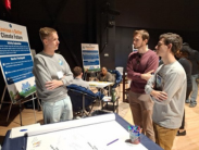 Three people stand in conversation near a display table at an event. Behind them are posters titled “Envision a Better Climate Future” 