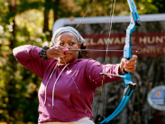 A woman in a maroon hoodie pulls back the string of a blue bow, aiming an arrow during an archery activity outdoors.