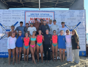 A group of lifeguards pose together on the beach in front of a banner.