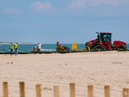 Workers and heavy equipment on a beach move large pipes as part of a sand bypassing project, with the ocean in the background.