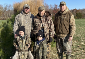 Four generations dressed in camouflage pose in a field on a sunny day. Two young boys in front hold geese while adults stand behind them.