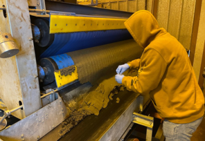 Person in a yellow hoodie and gloves collects a sample from wet material moving on a belt press inside an industrial facility.