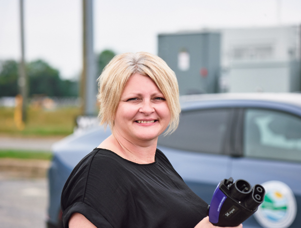 Breanne Preisen smiles while holding an electric vehicle charging plug near a DNREC EV at a public charging station.