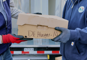 Two people wearing work gloves pass a cardboard box filled with venison labeled “DE Hunger-10” in front of a truck.