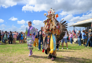 Two Nanticoke dancers in traditional regalia perform at an outdoor powwow in Milton as a large crowd looks on.