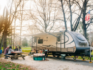 RV parked at a camp ground beside a fire pit and people sitting at a picnic table.