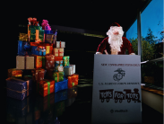 Santa poses beside a pile of wrapped gifts holding a Toys for Tots empty box.