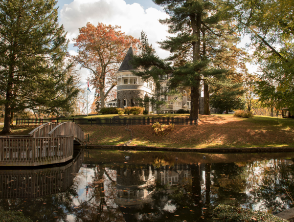 A stone mansion with a turret overlooks a pond and wooden footbridge, surrounded by fall trees reflected in the calm water.