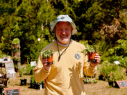 Man in a bucket hat and long sleeve yellow shirt smiles at the camera holding two small plants, in front of many plants for sale in the background.