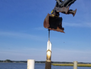 Construction bucket lowers a cylindrical object onto a dock piling over the water beneath a clear blue sky.