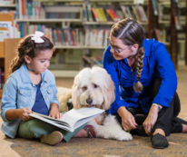 Young girl and woman reading to therapy dog in a library