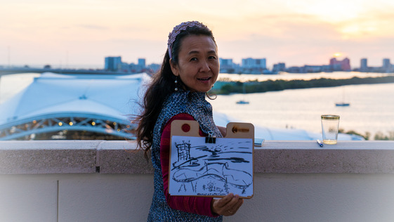 Image of a woman showing her drawing, standing in front of the sunset