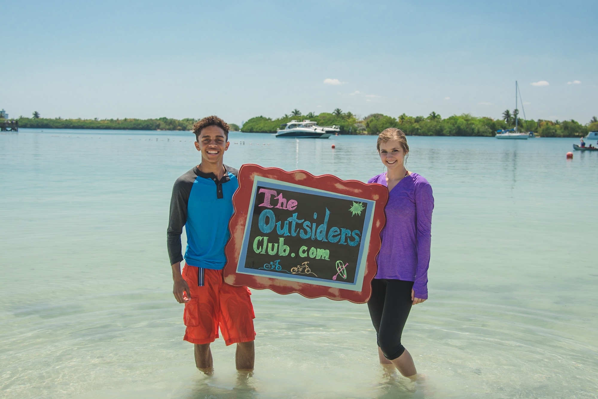 Two children stand in the water holding a sign that reads TheOutsidersClub.com
