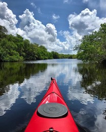 The front of a red kayak overlooks a river framed by trees and blue sky