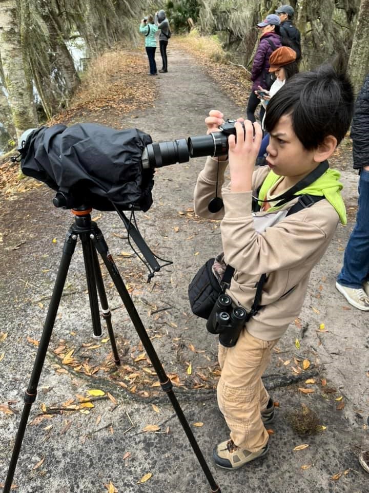 A young boy wearing brown and yellow stands on a dirt path looking through a black spotting scope. He has dark hair and a focused expression.