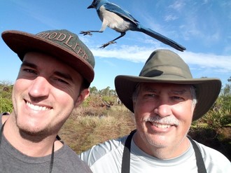 A father and son smiling in front of a scrub landscape and blue sky as a blue and white Florida Scrub-jay jumps between their hats.
