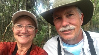 A close up of a smiling retired couple outdoors in Florida wearing hats, tshirts and binoculars.