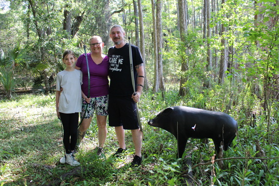 a family of three, smiling and holding bows, stands next to a foam target of a wild boar with an arrow in it, outside in the woods.