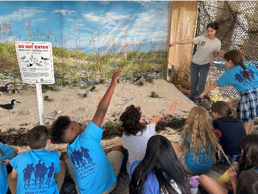 A group of elementary school students in front of a beach nest diorama while an FSA partner explains about shorebirds