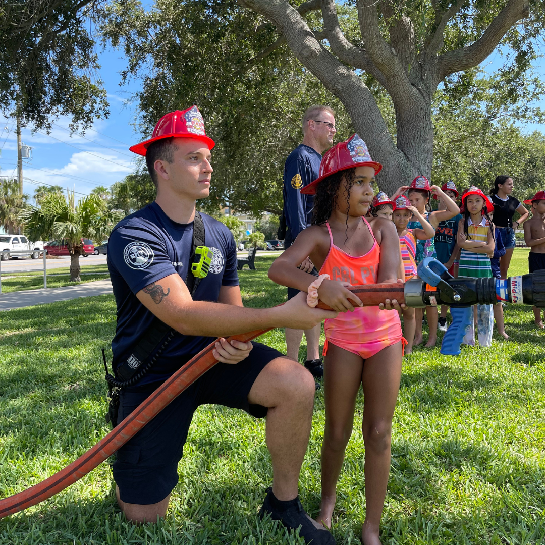 Treasure Island Fire Rescue Summer Camp Splash Down. Firefighter helping girl spray hose.