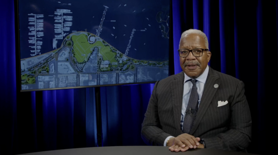 Mayor Keith James sits at desk with monitor in the background showing a West Palm Beach Waterfront rendering 