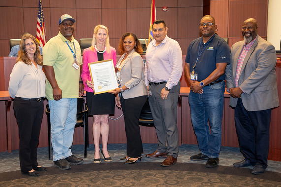 group of seven people stand together in a formal setting, with one person holding a framed certificate. 