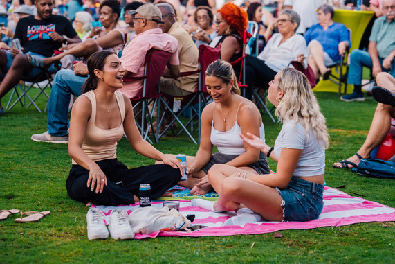 A group of people sitting on a pink striped blanket in a park, laughing and talking. 