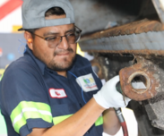 person wearing glasses and a cap is working on a vehicle part. They are holding a tool and wearing a uniform with a visible name tag. 