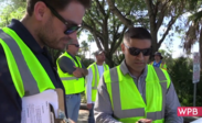 A group of people wearing reflective safety vests are outdoors. Two individuals in the foreground are focused on paperwork or a device. 